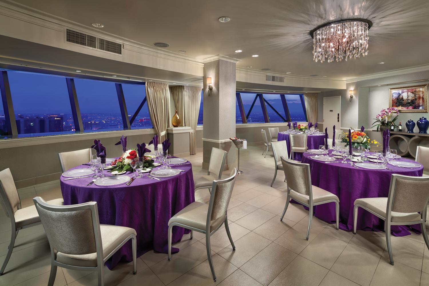 The STRAT Renaissance room in the SkyPod Tower with white tiles and a chandelier and tables with purple tablecloths set with dinnerware overlooking a view of the Las Vegas valley